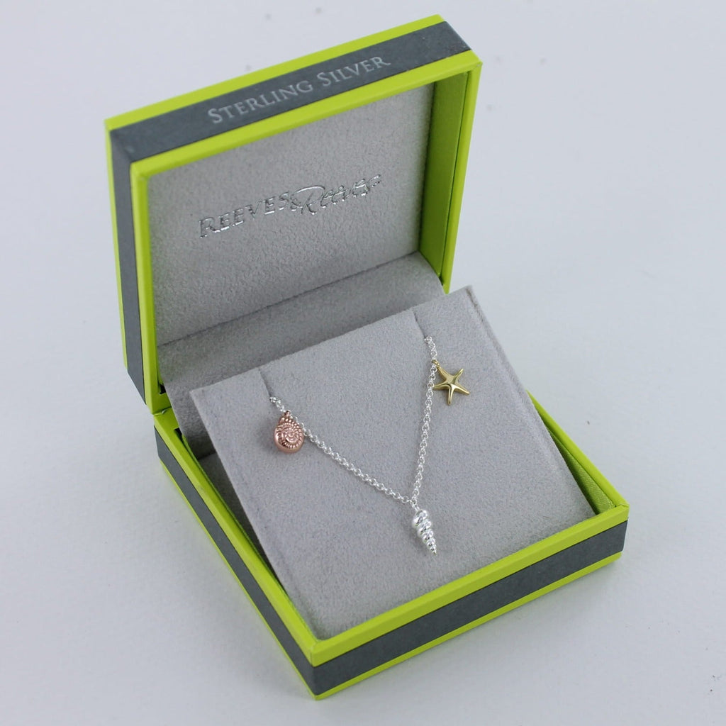 A silver chain necklace with a silver conch shell, rose gold plated ammonite shell and yellow gold plated starfish. Shown in a Reeves & Reeves grey and green branded gift box.