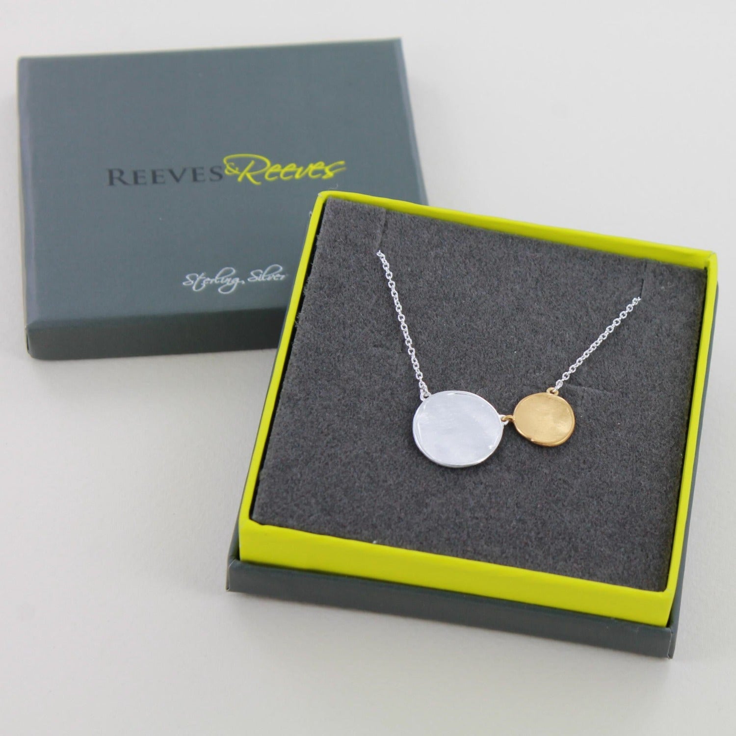 A Silver and gold plate disc necklace, one silver disc and one gold plated disc sit next to each other on the silver chain and shown in a Reeves & Reeves grey and green gift box.