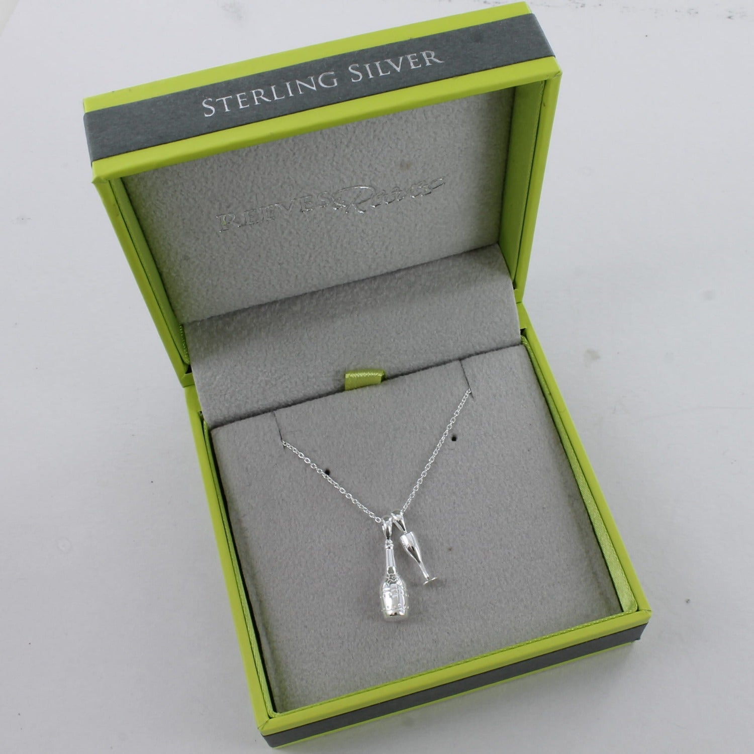 Sterling Silver champagne bottle with a glass charm, sat next to each other on a silver chain and shown in a Reeves & Reeves grey and green gift box.