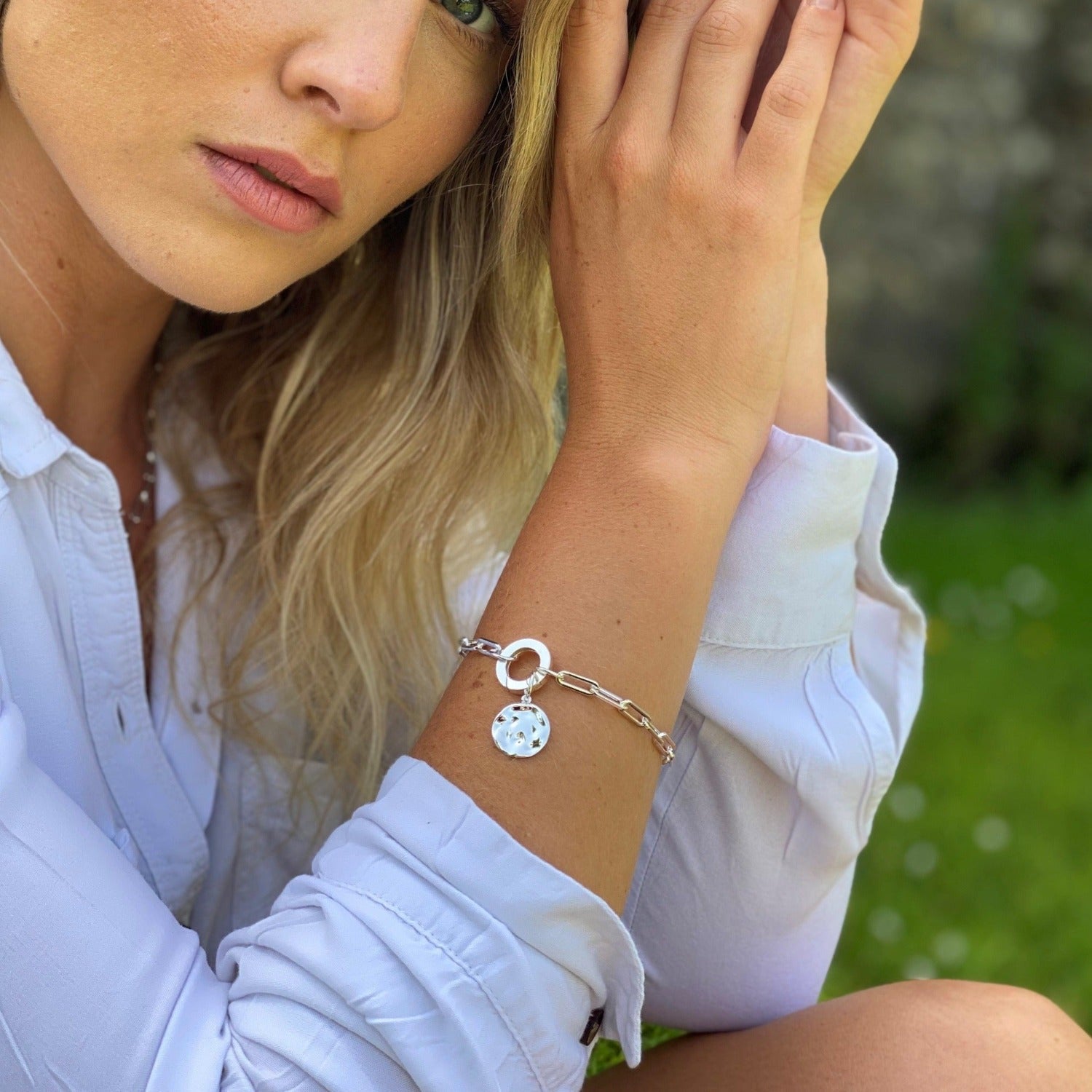 Silver paperclip style chain bracelet with a circular clasp and a hammered disc charm hanging from the bracelet. Worn on a women's wrist.