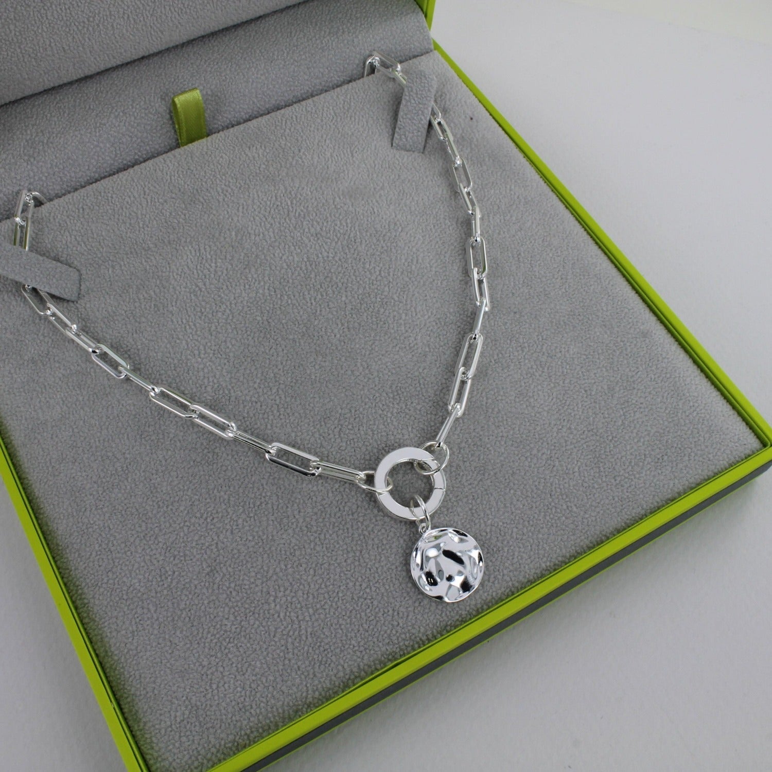 A silver paperclip style chain with a toggle clasp and a hammered disc charm hanging below. Shown in a Reeves & Reeves branded grey and green gift box.