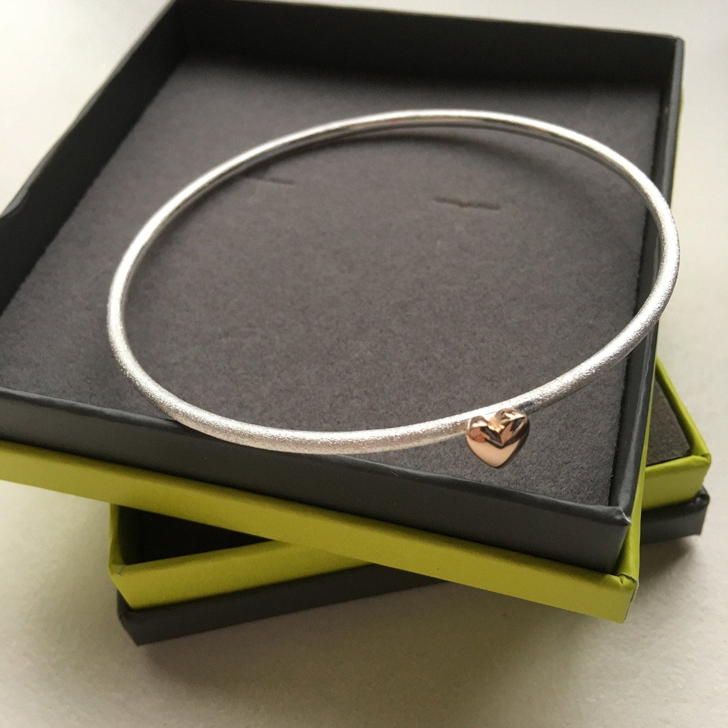 A Silver matt finish bangle with a rose gold high shine heart attached, shown in a Reeves & Reeves branded grey and green gift box.