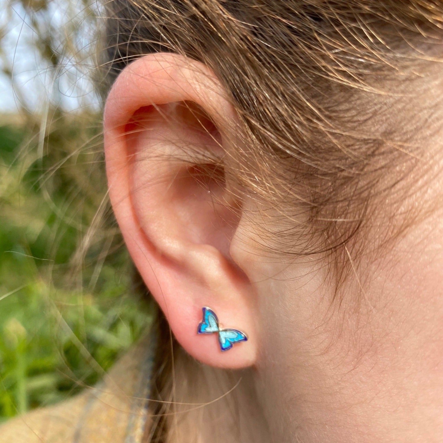 A pair of sterling silver Butterfly stud earrings with blue enamel detail, shown in a ladies ear.