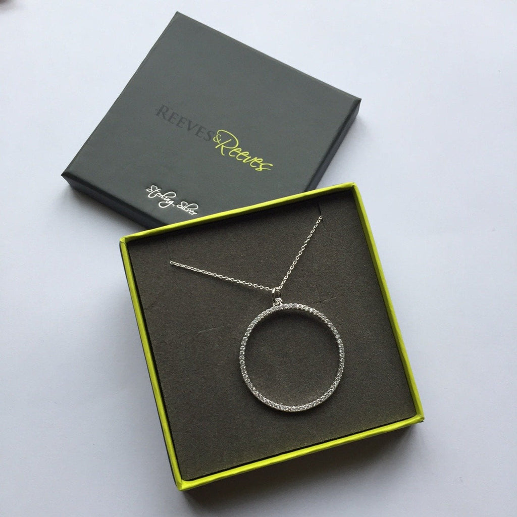 A sterling silver circular pendant, filled with cubic zirconia stones and hung on a silver adjustable chain. Shown in a Reeves & Reeves grey and green branded gift box.