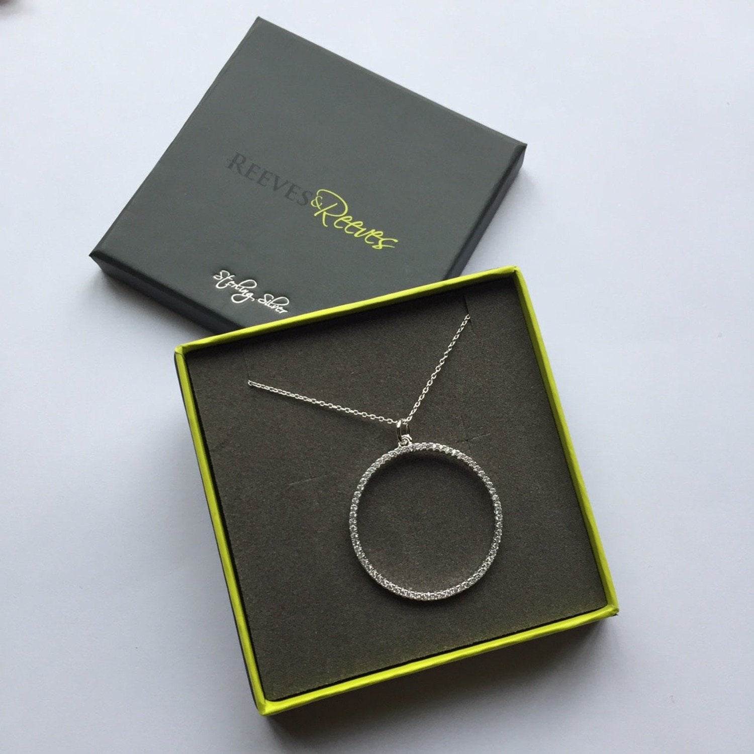 A sterling silver circular pendant, filled with cubic zirconia stones and hung on a silver adjustable chain. Shown in a Reeves & Reeves grey and green branded gift box.