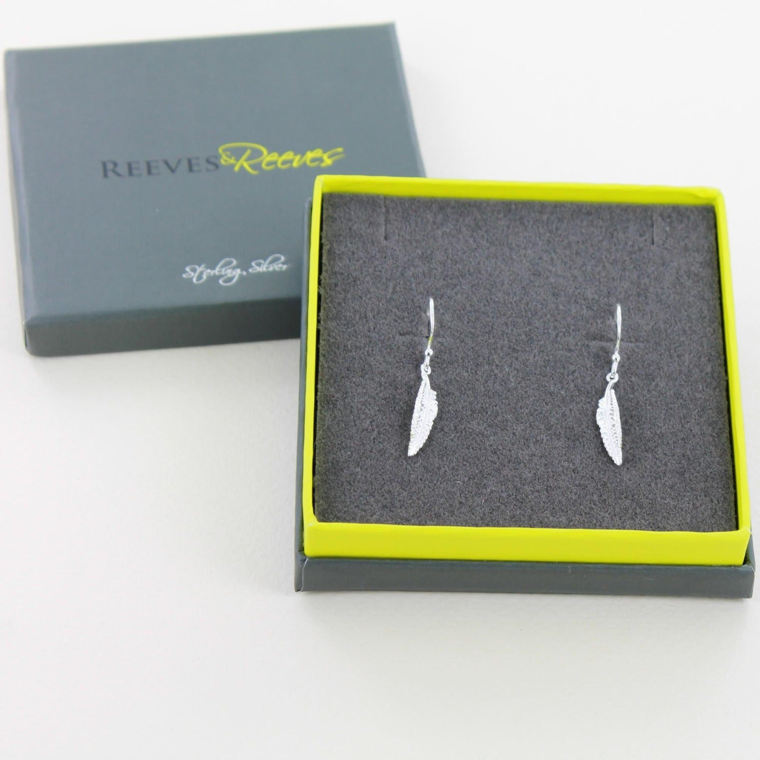 A pair of sterling silver feather drop earrings, hung on silver hooks and shown in a Reeves & Reeves grey and green gift box.