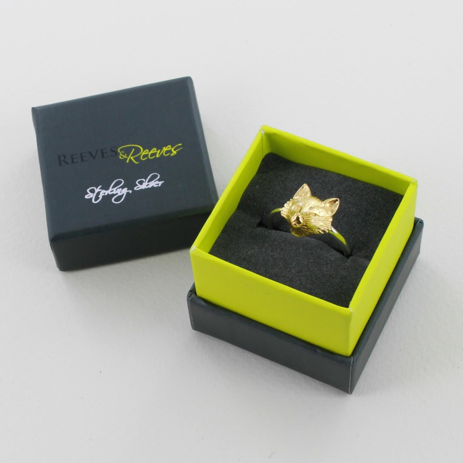 A silver banded adjustable ring with a gold plated fox head, shown in a Reeves & Reeves grey and green branded gift box.