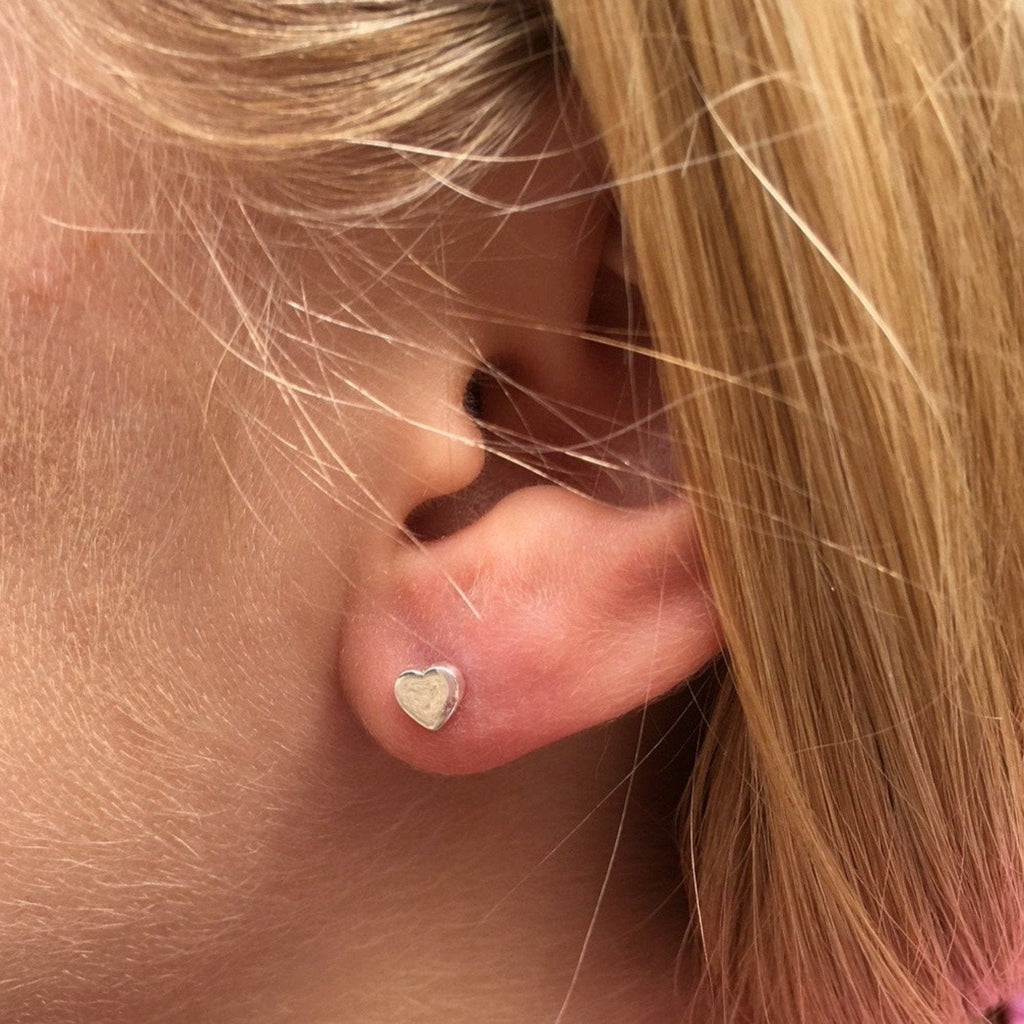 A pair of silver heart studs, three dimensional in style and shown in a ladies ear.