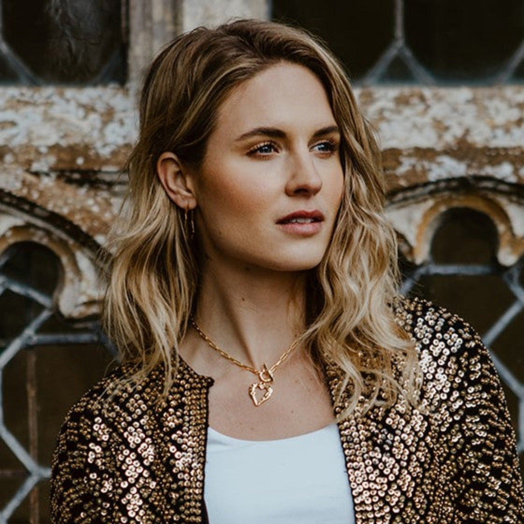 A sterling silver and gold plated silhouette heart pendant with a hammered finish. The heart hangs from a circular clasp with a t bar and on a paperclip style chain. Worn on a models neck.