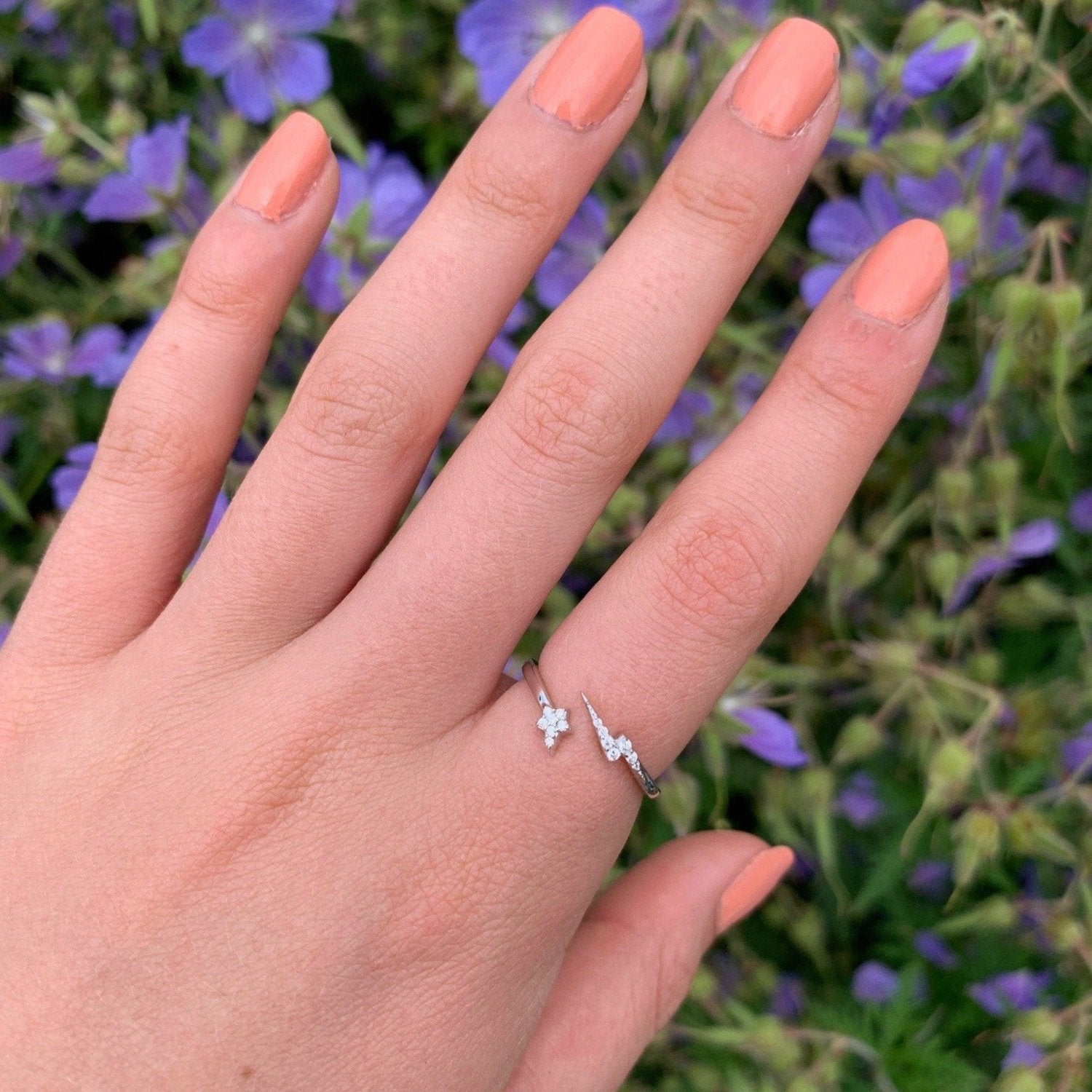 A Silver adjustable banded ring with a star at one end and a lightening bolt at the other, both filled with cubic zirconia stones. Worn on a women's hand with blue flowers in the background.