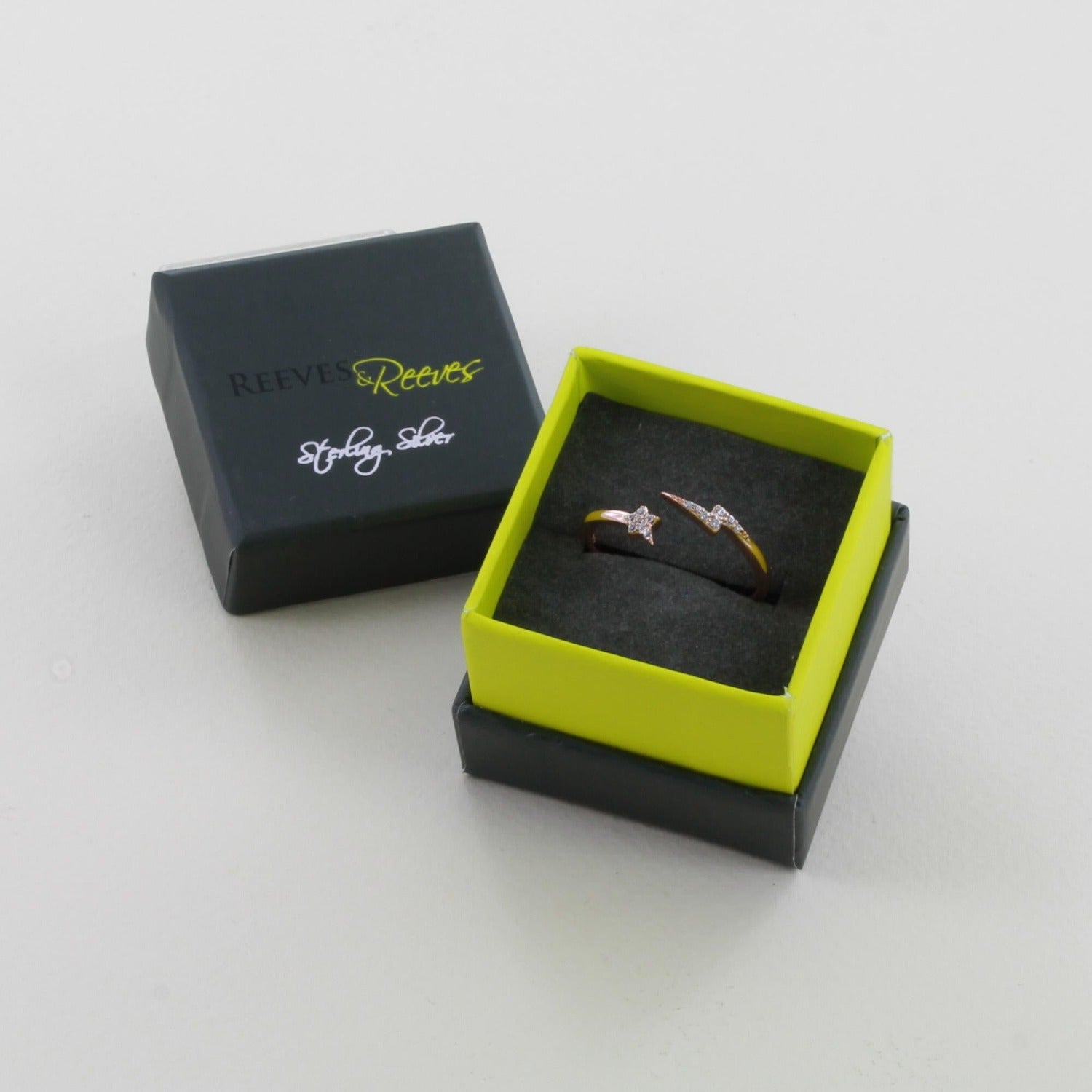 An adjustable banded rose gold ring with a star at one end and a lightening bolt at the other, shown in a Reeves & Reeves grey and green branded gift box.