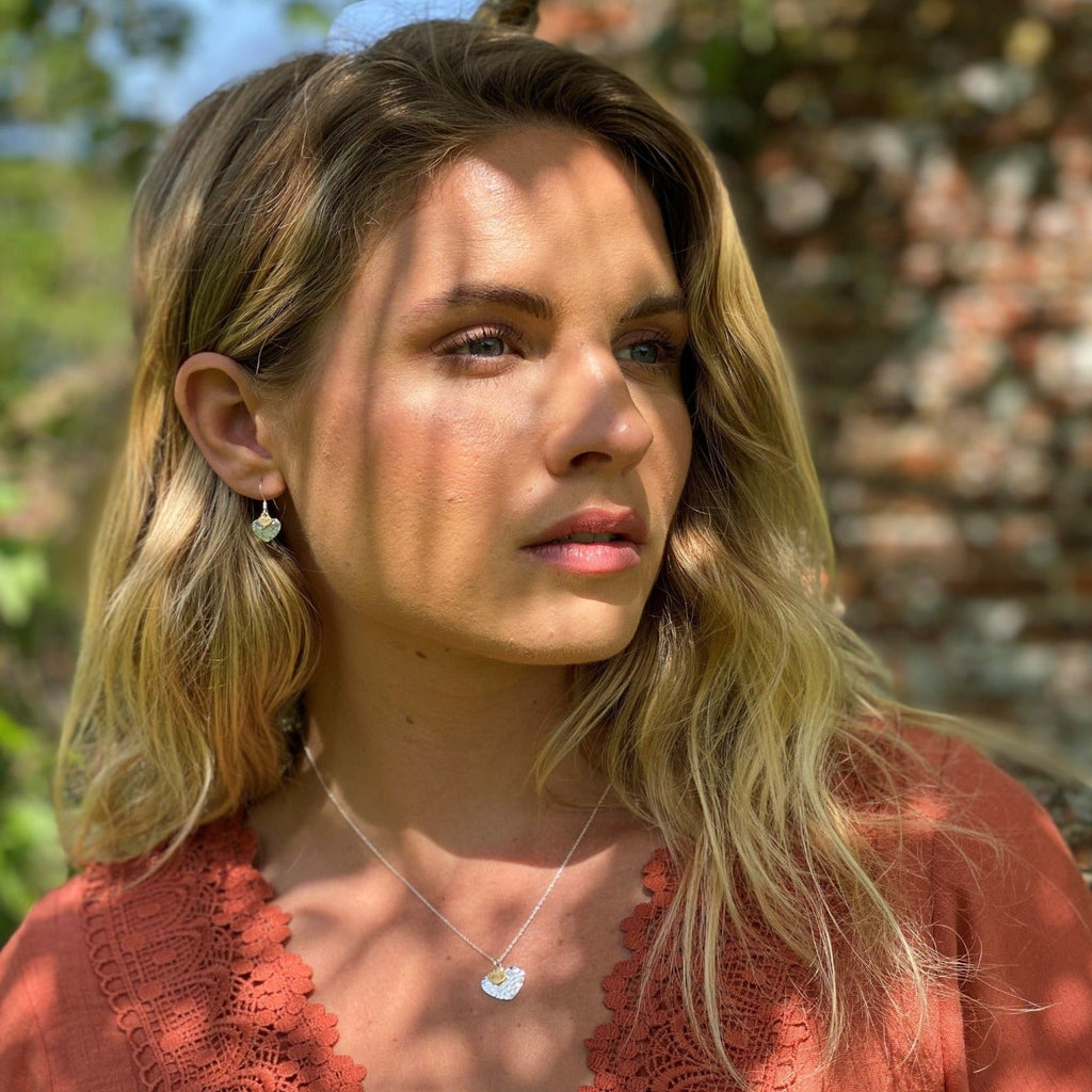 A hammered heart pendant with a smaller gold plated heart sat on top with the word 'love' enscribed and worn on a model with the matching drop earrings.