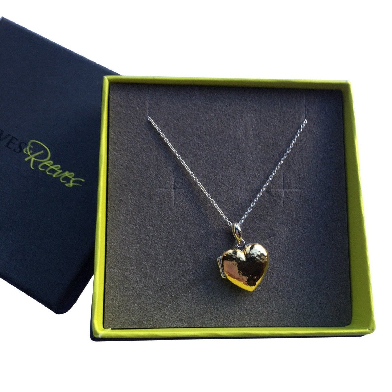 A heart shaped locket with a hammered texture, hanging on a silver adjustable chain and in a grey and green branded gift box.
