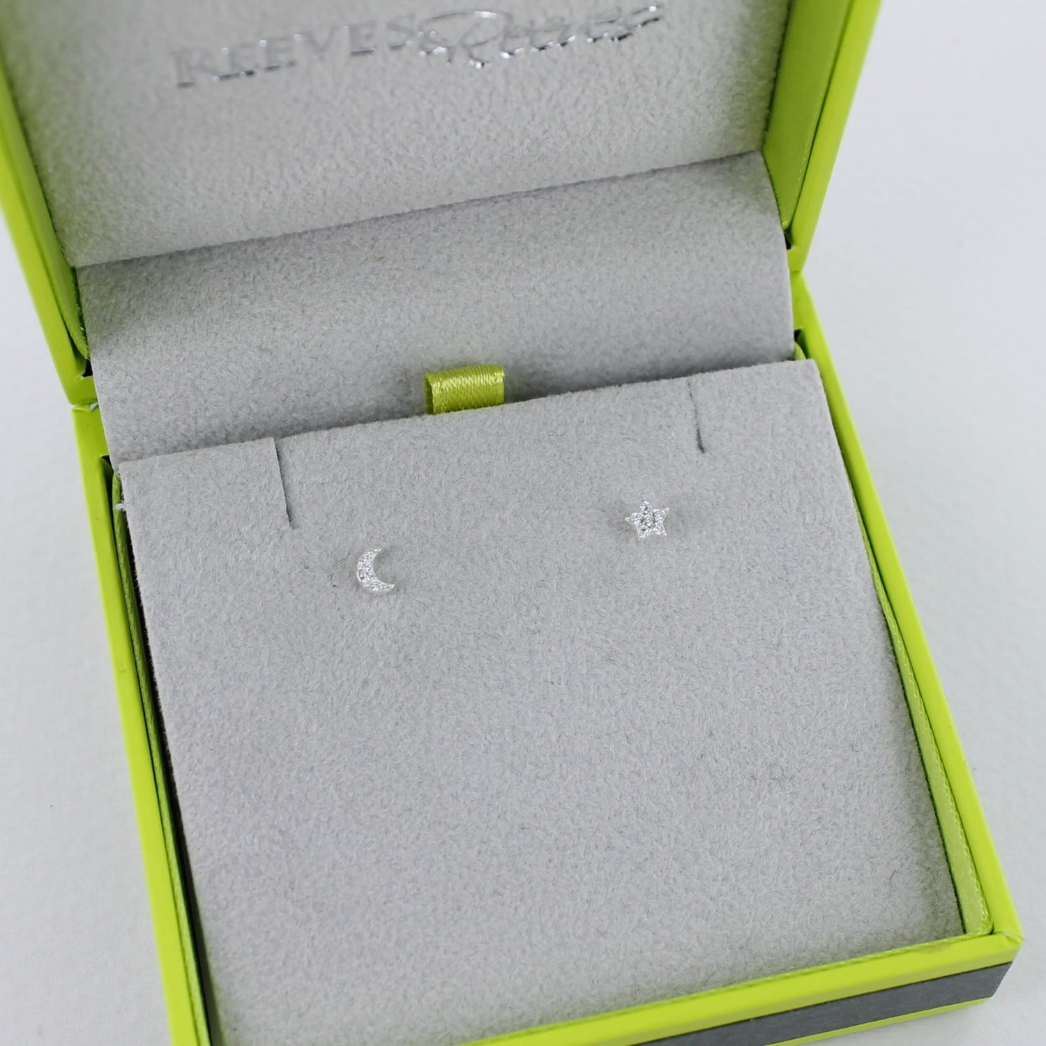 A pair of sterling silver mini moon and star stud earrings, shown in a Reeves & Reeves grey and green gift box.