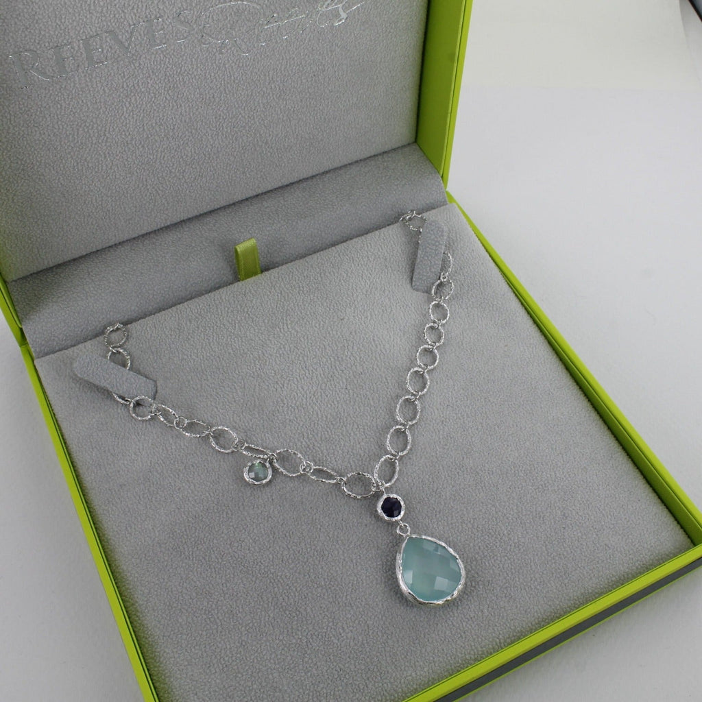A sterling silver oval paper link style chain with textured finish on the ovals. Hanging from the centre is an amethyst stone, and below a larger peardrop aqua chalcedony stone. There is also a moonstone hanging further up the chain. Shown in a Reeves & Reeves gift box.