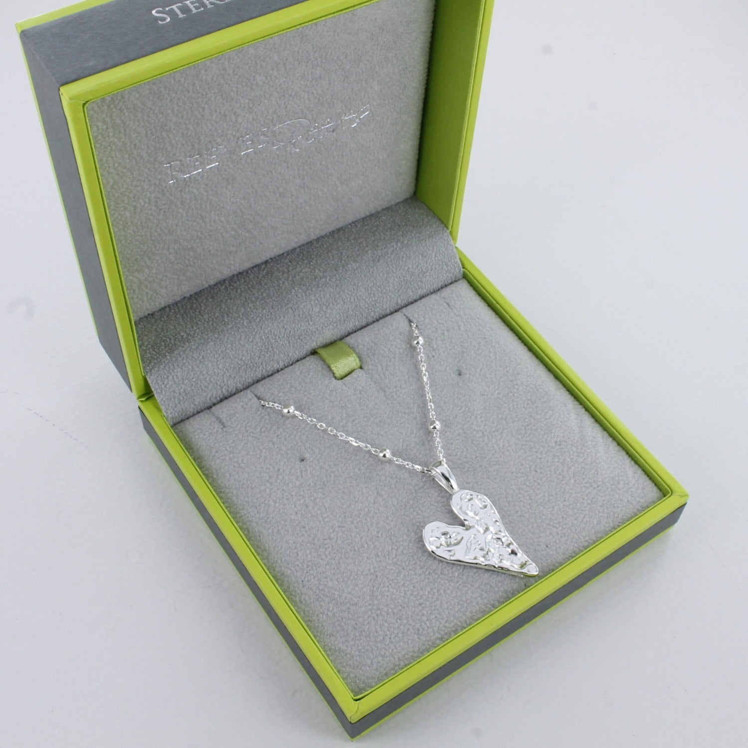 Solid silver textured heart hanging on a silver adjustable beaded chain, shown in a Reeves & Reeves branded grey and green gift box.