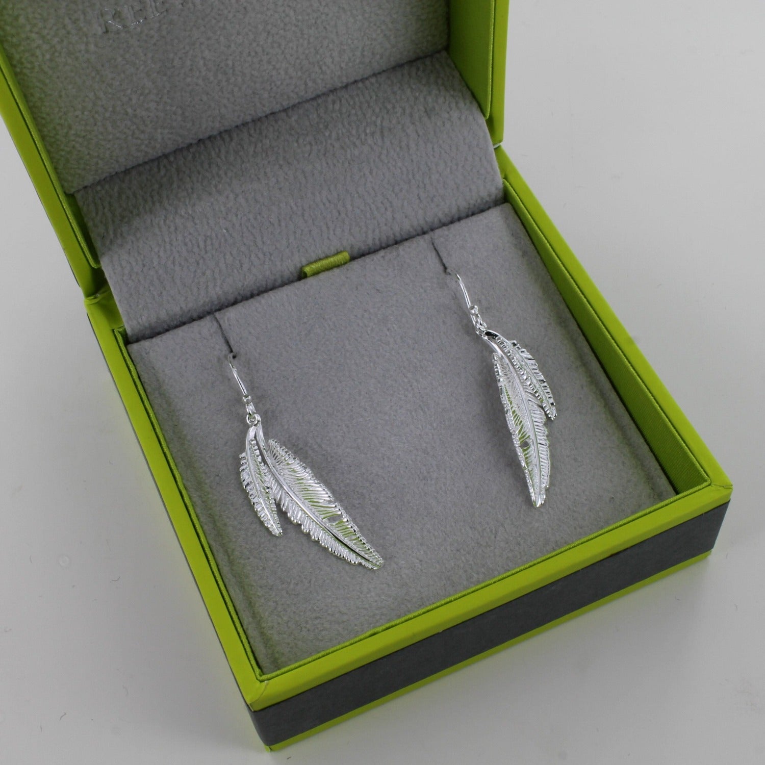 A pair of sterling silver feather drop earrings, two feathers of different sizes sit next to each other and hang on silver hooks. Shown in a Reeves & Reeves gift box.