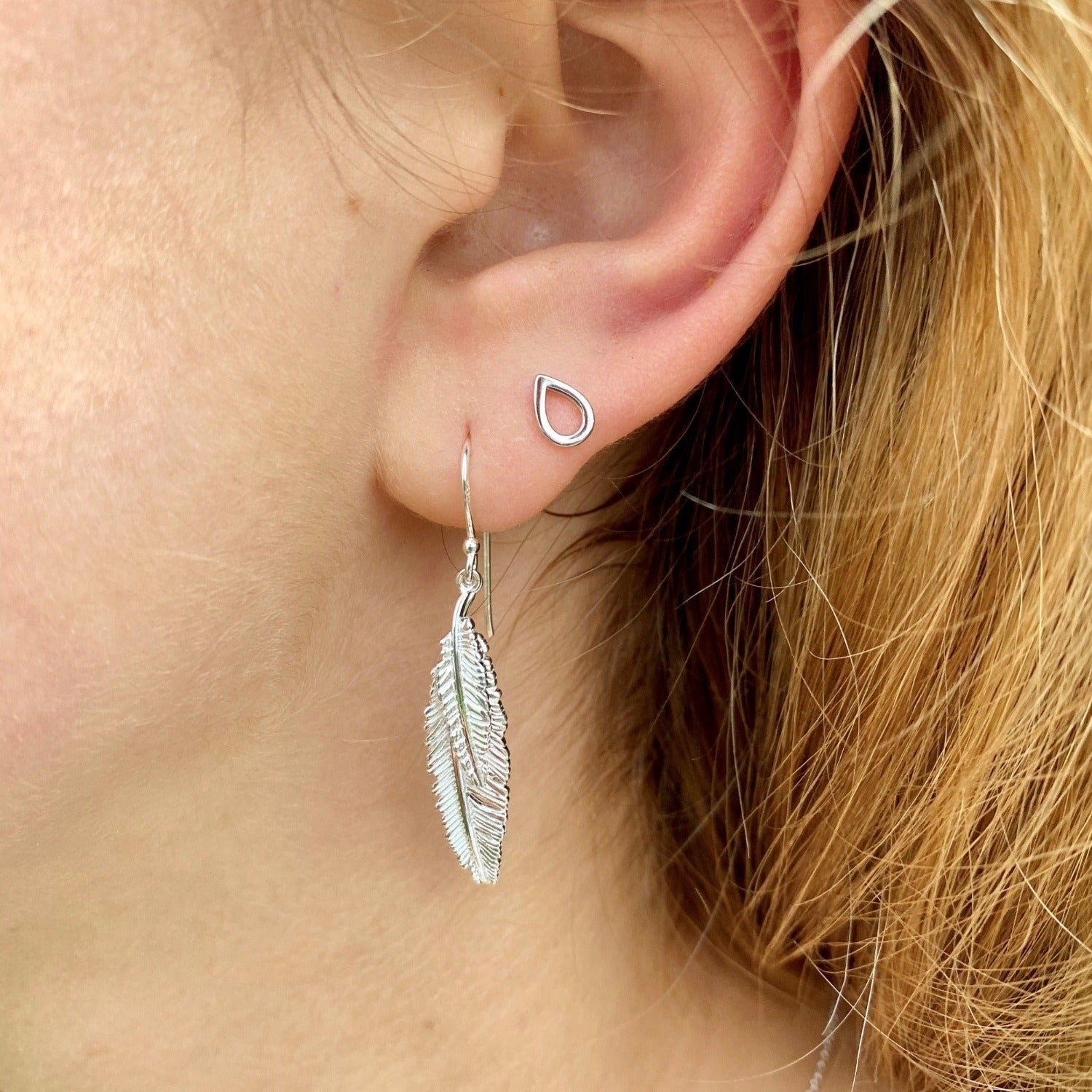 A sterling silver drop style earring, with two feathers, one smaller hung in front of the larger, hanging from silver hooks and shown in a ladies ear.