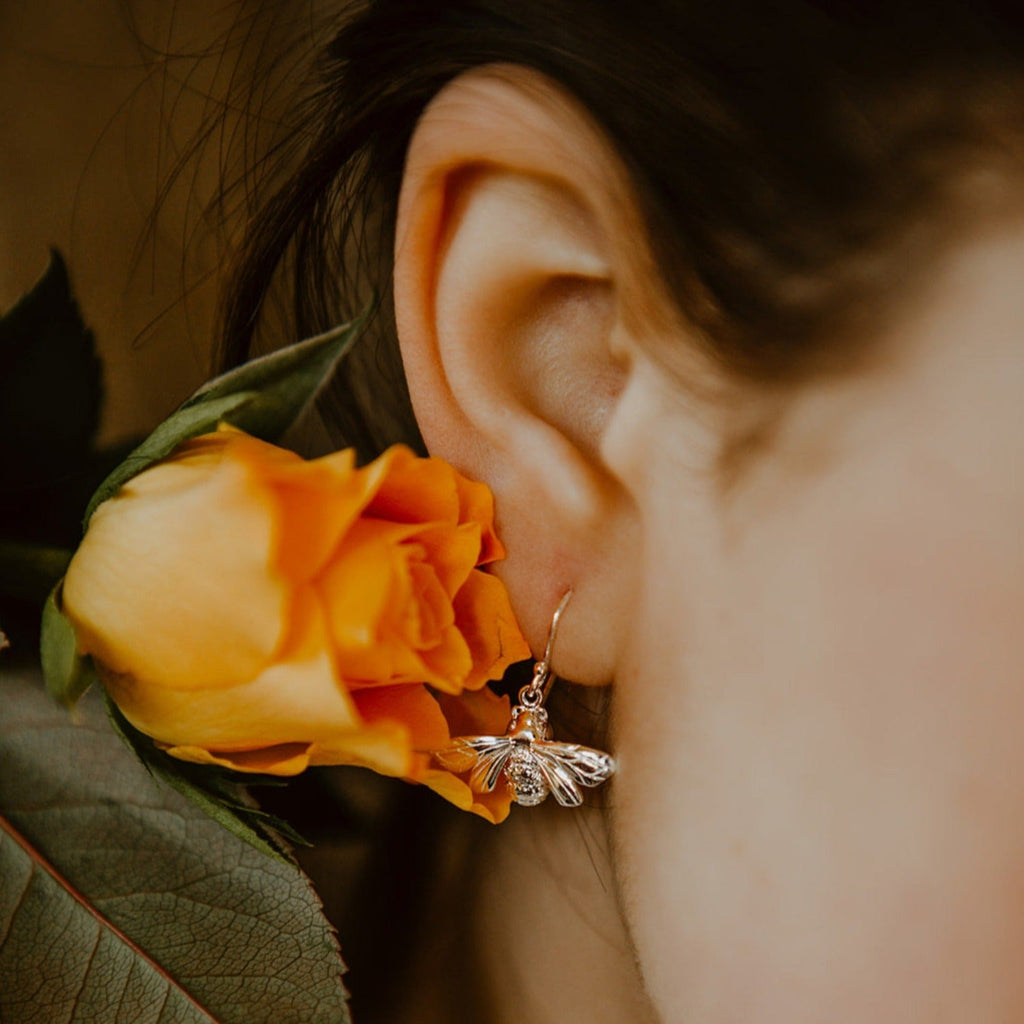 A sterling silver Queen Bee Drop Earring, hung on a silver hook and shown in a ladies ear next to an orange rose.