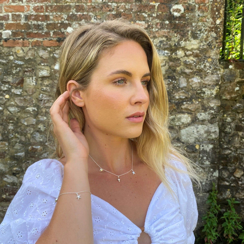 A silver rockpool necklace showing a starfish, scallop and conch shell charm hanging separately around the chain. Worn on a model with the matching bracelet.