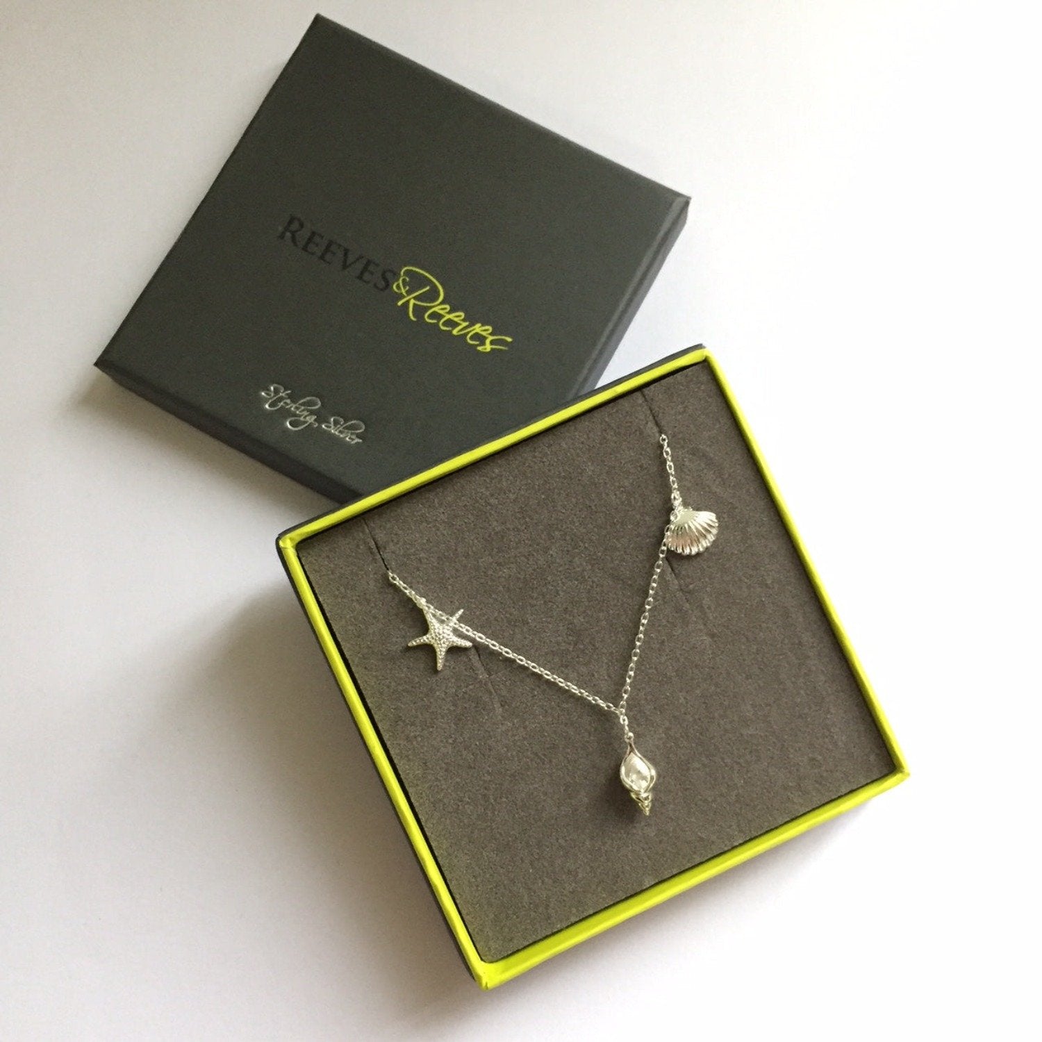 A silver rockpool necklace with three beach charms, a starfish, conch and scallop shell charms. Shown in a Reeves & Reeves grey and green branded gift box.