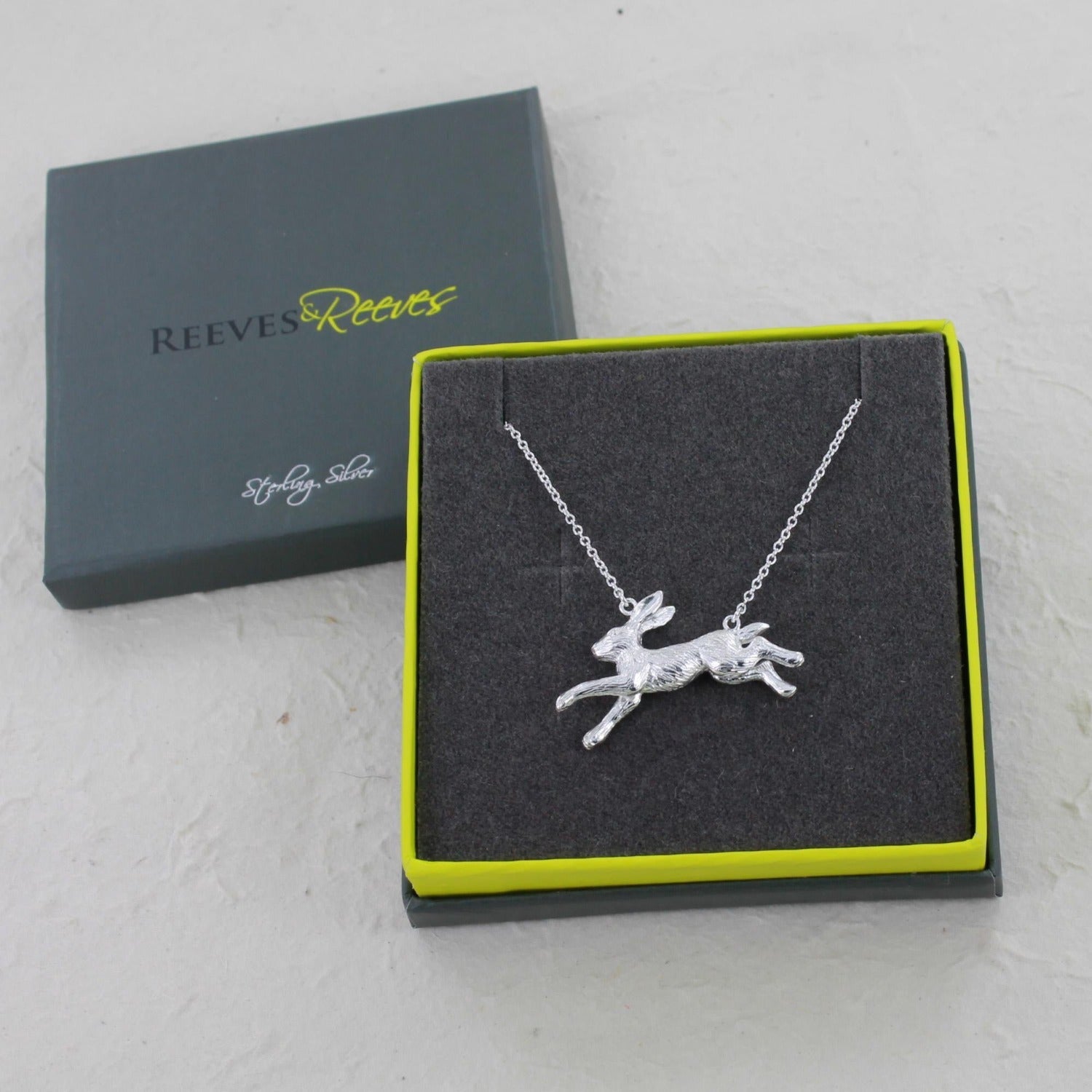 A Silver Running Hare Necklace, hanging on an adjustable silver chain, shown in a Reeves & Reeves branded gift box in grey and green colours.