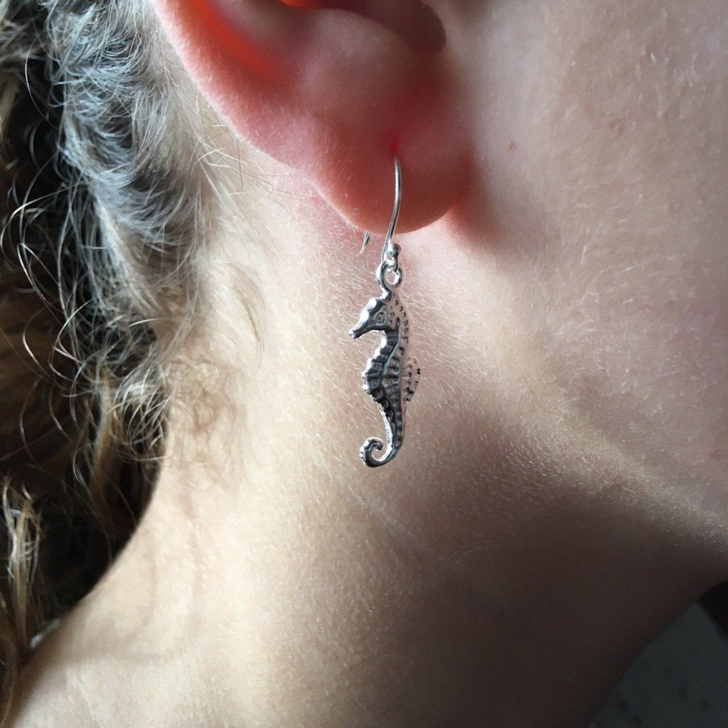 A sterling silver seahorse charm, hanging on the bottom of a silver hook to create these drop earrings shown in a ladies ear.