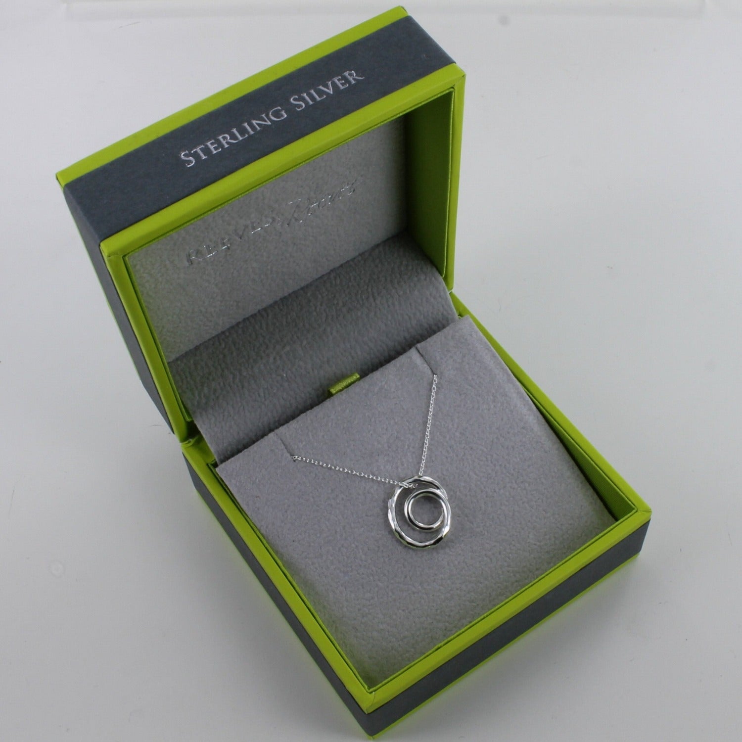 A circular 'sending hugs' necklace, shown in a Reeves & Reeves grey and green branded gift box.