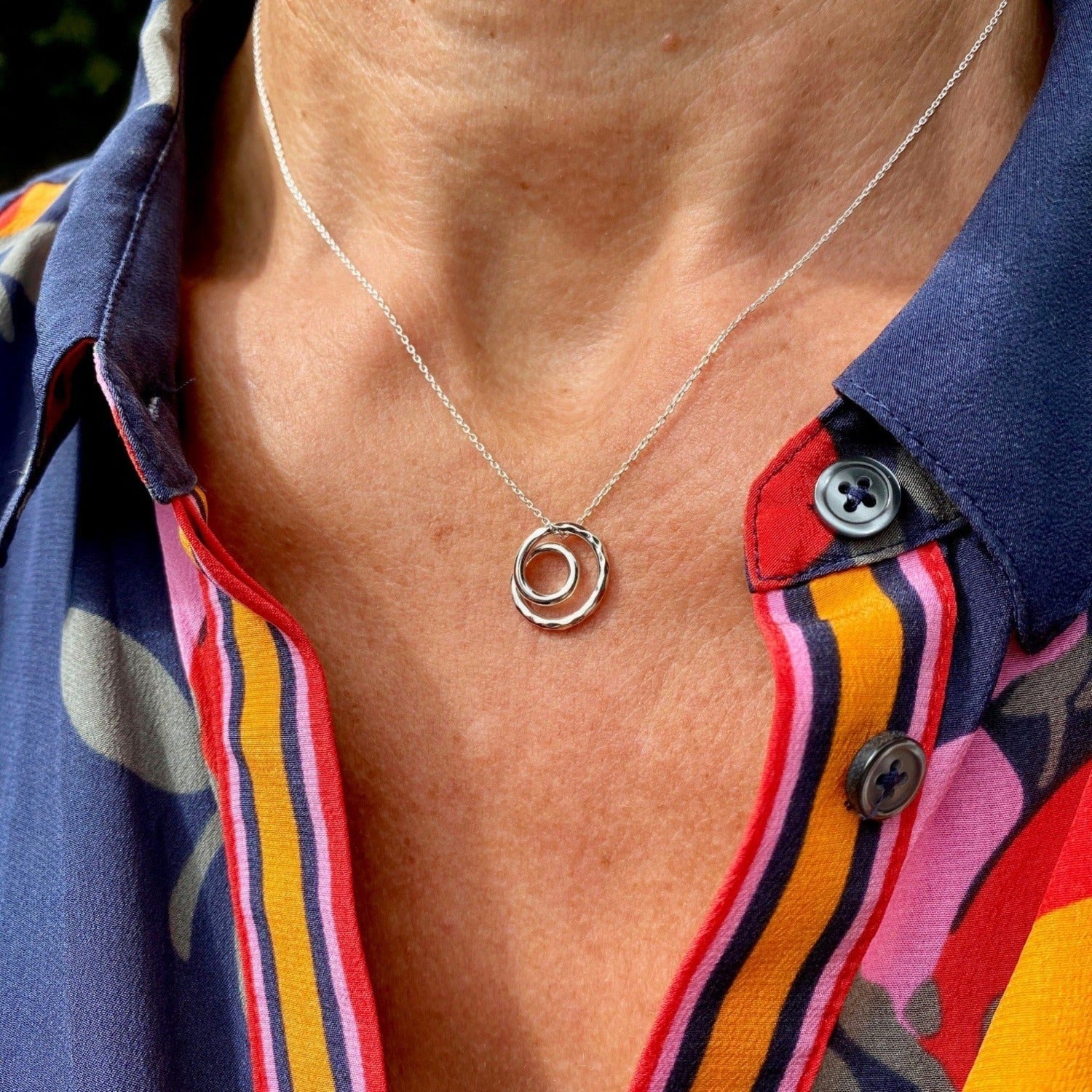 A silver circular pendant with hammered finish worn on a ladies neck with an open blouse.