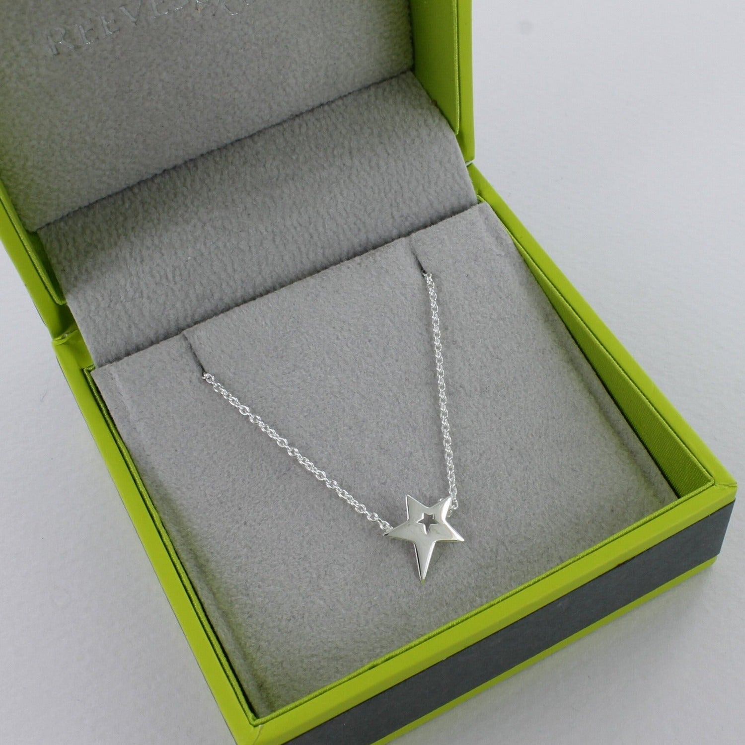 Silver star necklace with a cut out centre, hanging on a silver adjustable chain and in a Reeves & Reeves grey and green branded gift box.