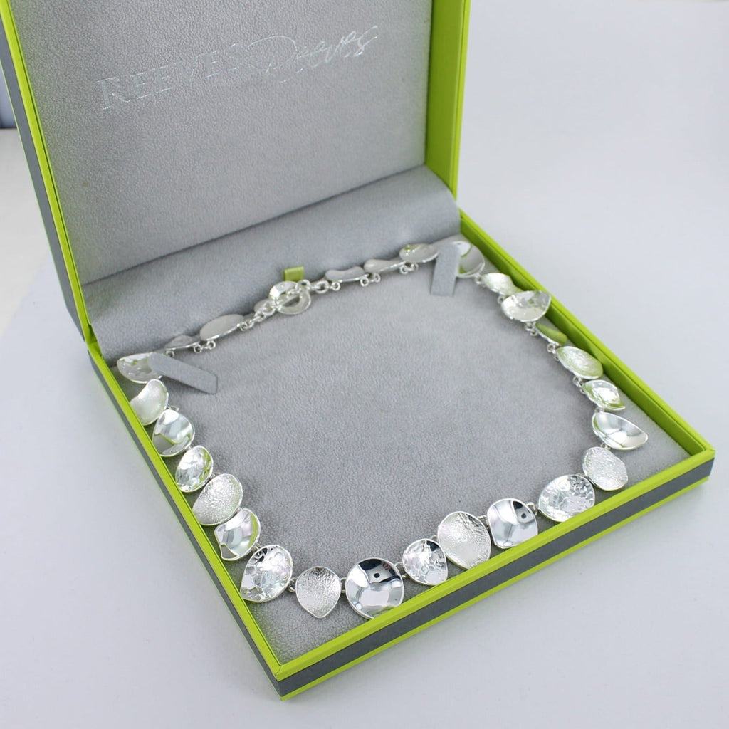 A sterling silver necklace comprising of irregular circular shapes of varying textures, finished with a t bar clasp and shown in a Reeves & Reeves gift box.