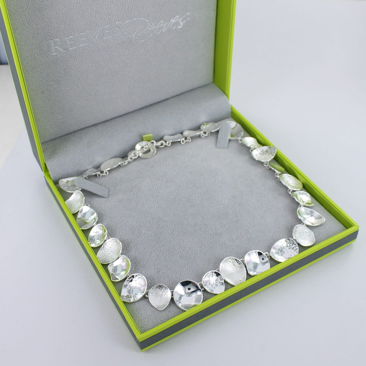 A sterling silver necklace comprising of irregular circular shapes of varying textures, finished with a t bar clasp and shown in a Reeves & Reeves gift box.