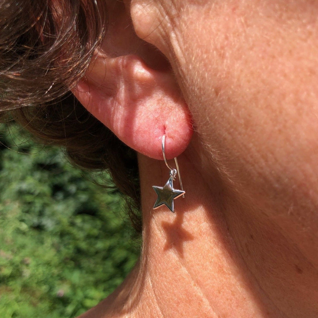 A pair of sterling silver star drop earrings, the flat star charms hang from silver hooks, worn in a ladies ear.