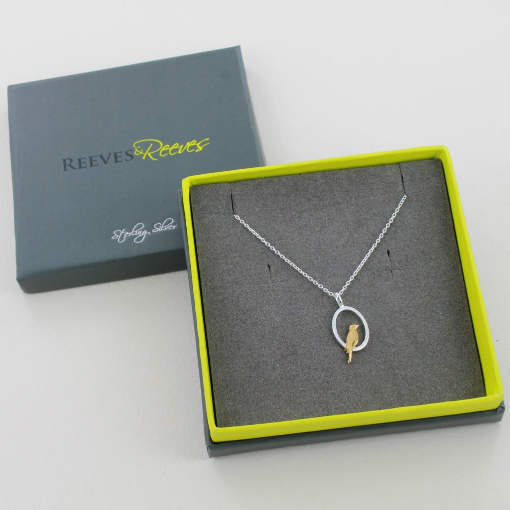 A silver oval pendant with a gold plate bird sat inside and hanging on a silver adjustable chain. Shown in a Reeves & Reeves gift box.