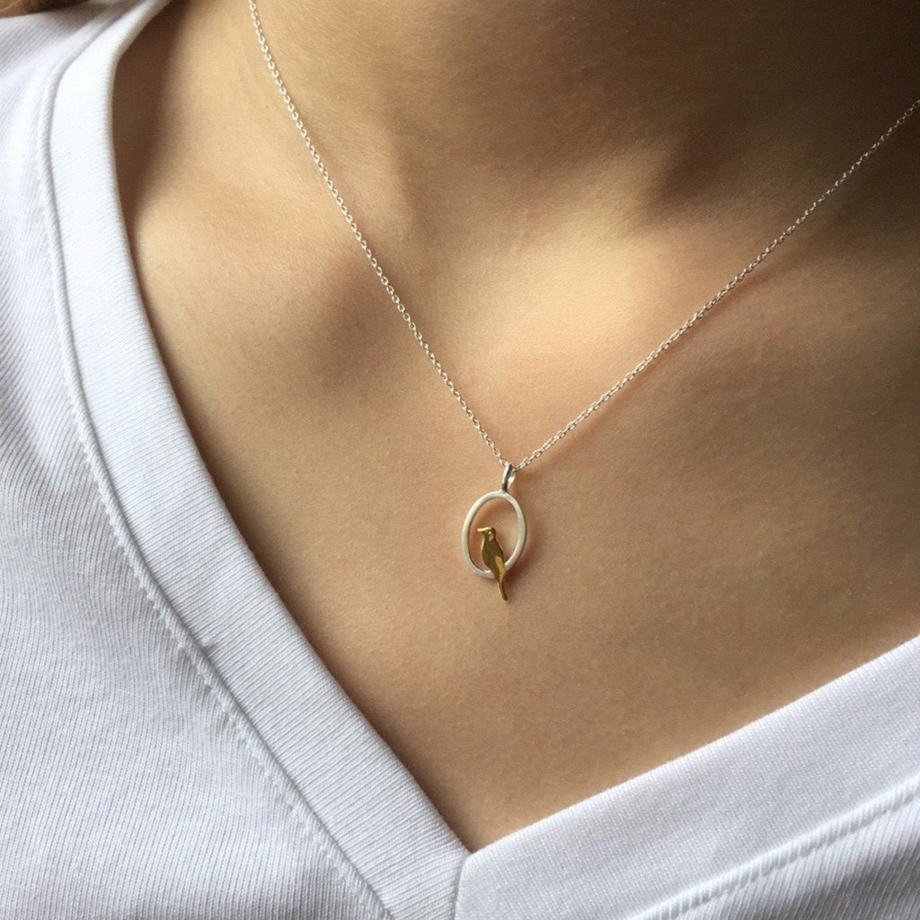 A silver oval pendant with a gold plated bird sat inside, hanging on a silver chain and worn on a models neck.