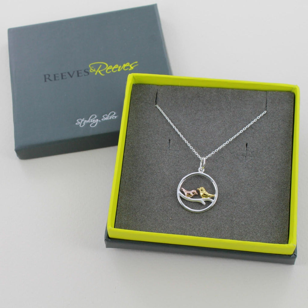 A sterling silver circular pendant with two birds sat on a branch inside, one gold plate and one rose gold plate on a silver chain. Shown in a Reeves & Reeves branded gift box.