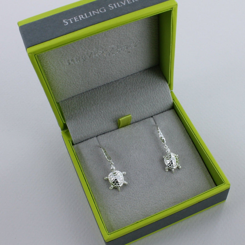 A pair of Turtle Drop Earrings, hanging on hammered hooks and shown in a Reeves & Reeves grey and green gift box.