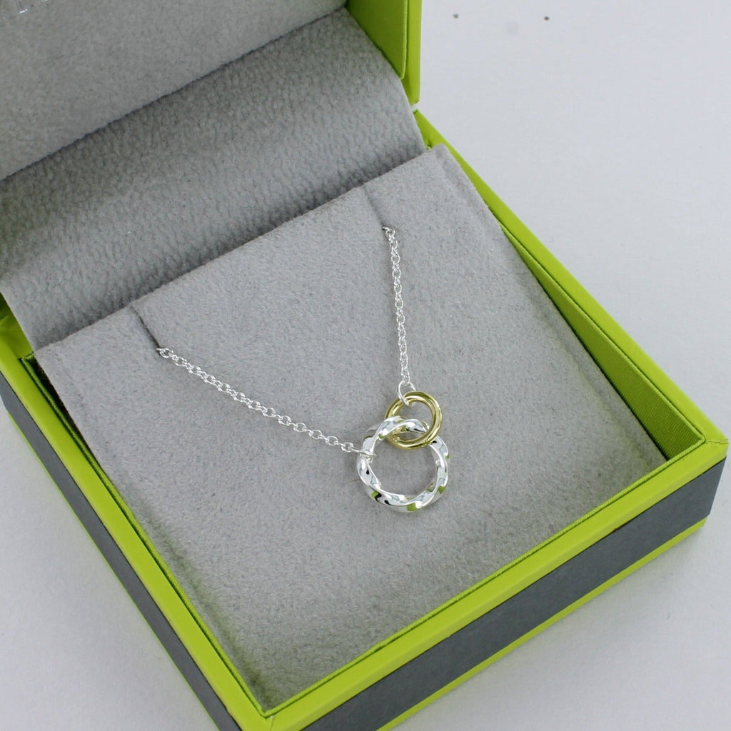 A silver twisted open circle with a gold plated smaller circle interlinked and both hanging on a silver chain. Shown in a Reeves & Reeves grey and green branded gift box.