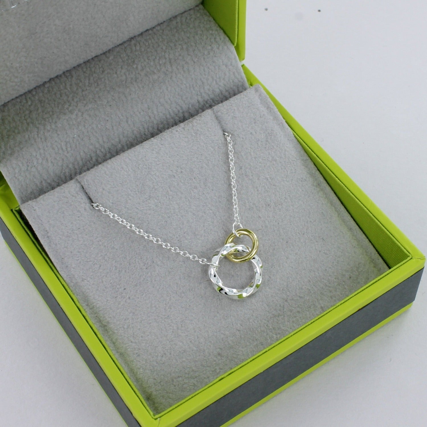 A silver twisted open circle with a gold plated smaller circle interlinked and both hanging on a silver chain. Shown in a Reeves & Reeves grey and green branded gift box.