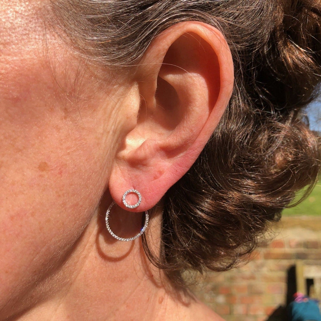A sterling silver earring which has a large circular shape filled with cubic zirconia stones that sits behind the ear, with a smaller circle, also filled with cubic zirconia sitting on the lobe. Shown in a ladies ear.