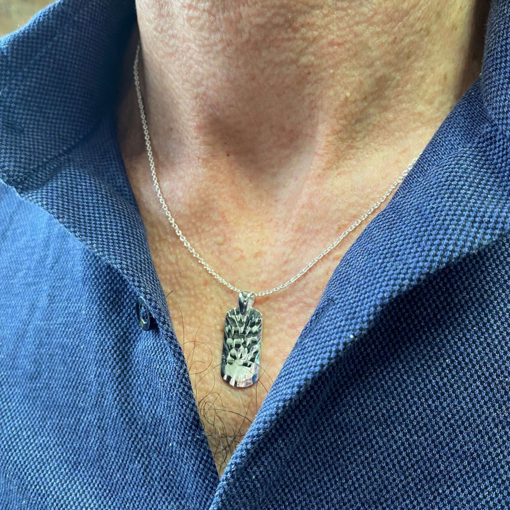 A textured dog tag pendant, rectangular in shape with slightly rounded edges and hung on a silver adjustable chain and worn on a man's neck.