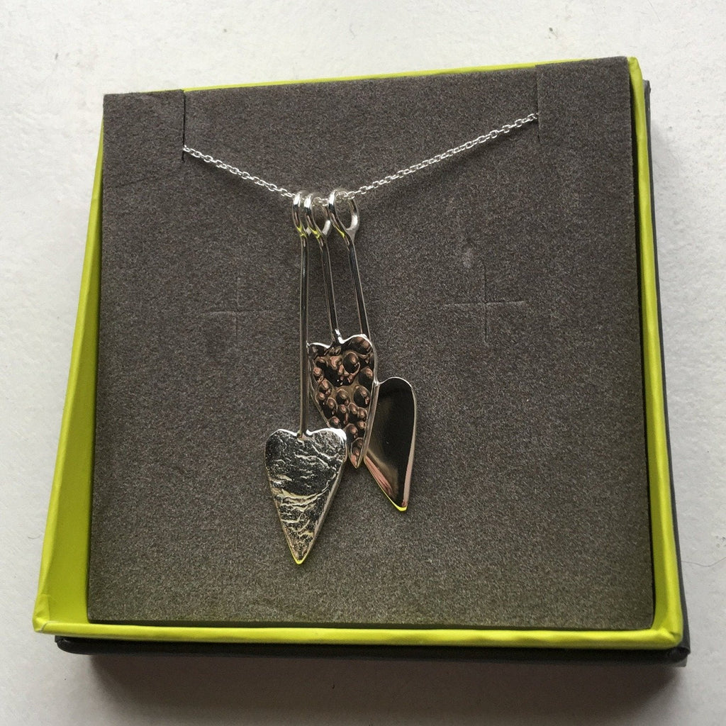 Three silver hearts with varying textures an hanging on silver hooks at varying lengths on a silver chain. Shown in a Reeves & Reeves branded gift box.