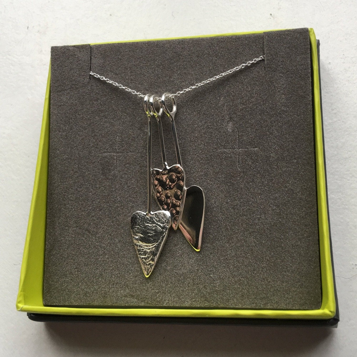 Three silver hearts with varying textures an hanging on silver hooks at varying lengths on a silver chain. Shown in a Reeves & Reeves branded gift box.