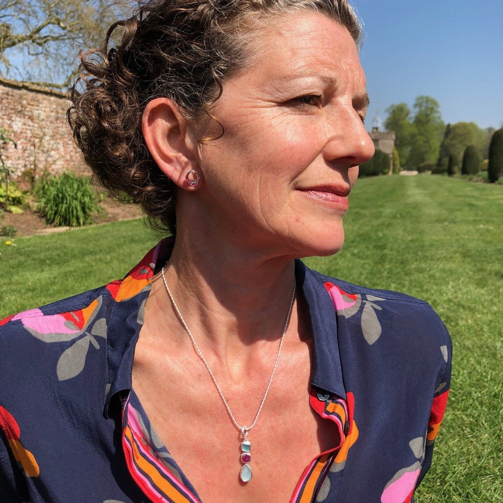 A sterling silver pendant with three rough cut stones, one apatite, one ruby and one aqua chalcedony in a vertical design, hung on a silver rope style chain and worn on a model.
