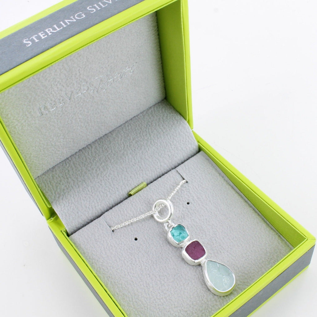 A sterling silver pendant with three rough cut stones, one apatite, one ruby and one aqua chalcedony, hung in a vertical design on a silver chain and shown in a Reeves & Reeves gift box.