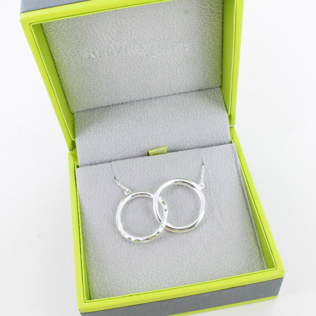 A silver twin ring necklace, with two interlocking rings one with a hammered finish and on a silver chain. Shown in a Reeves & Reeves grey and green gift box.