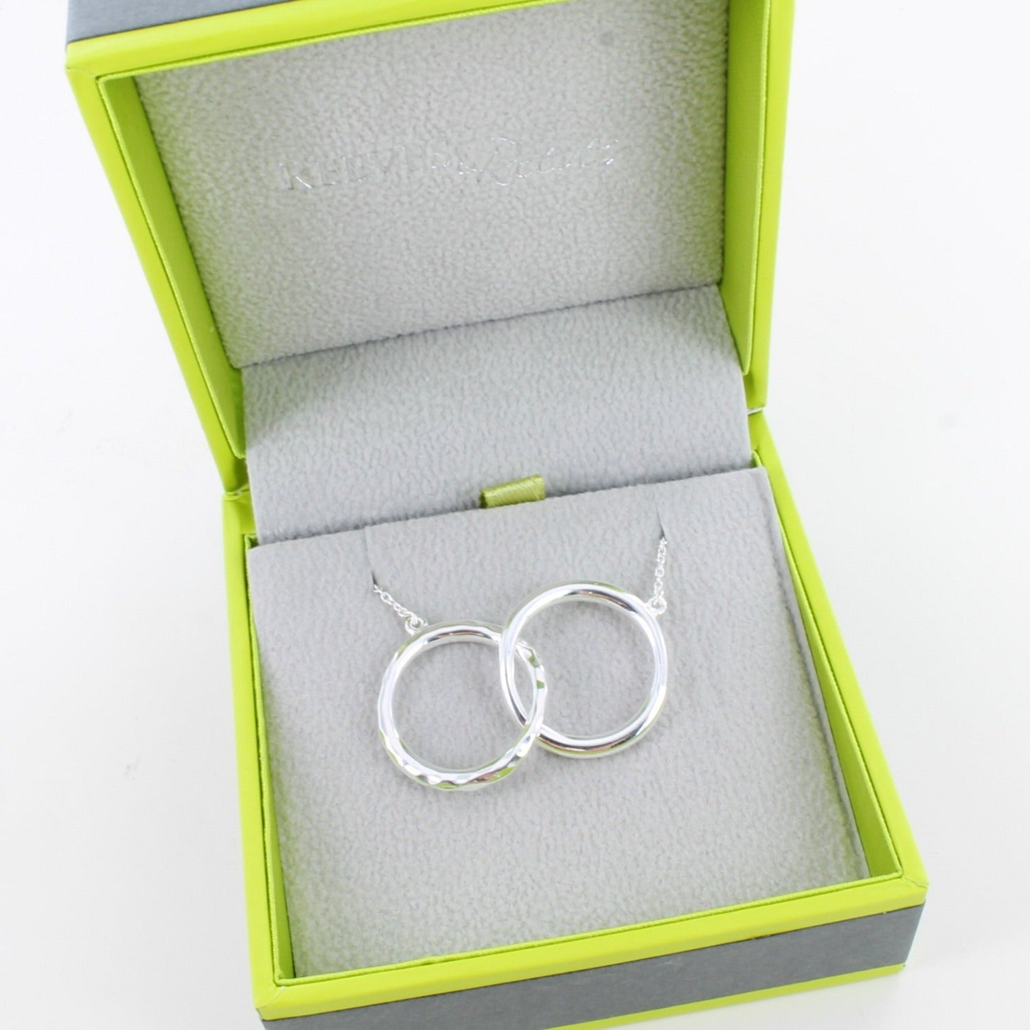 A silver twin ring necklace, with two interlocking rings one with a hammered finish and on a silver chain. Shown in a Reeves & Reeves grey and green gift box.